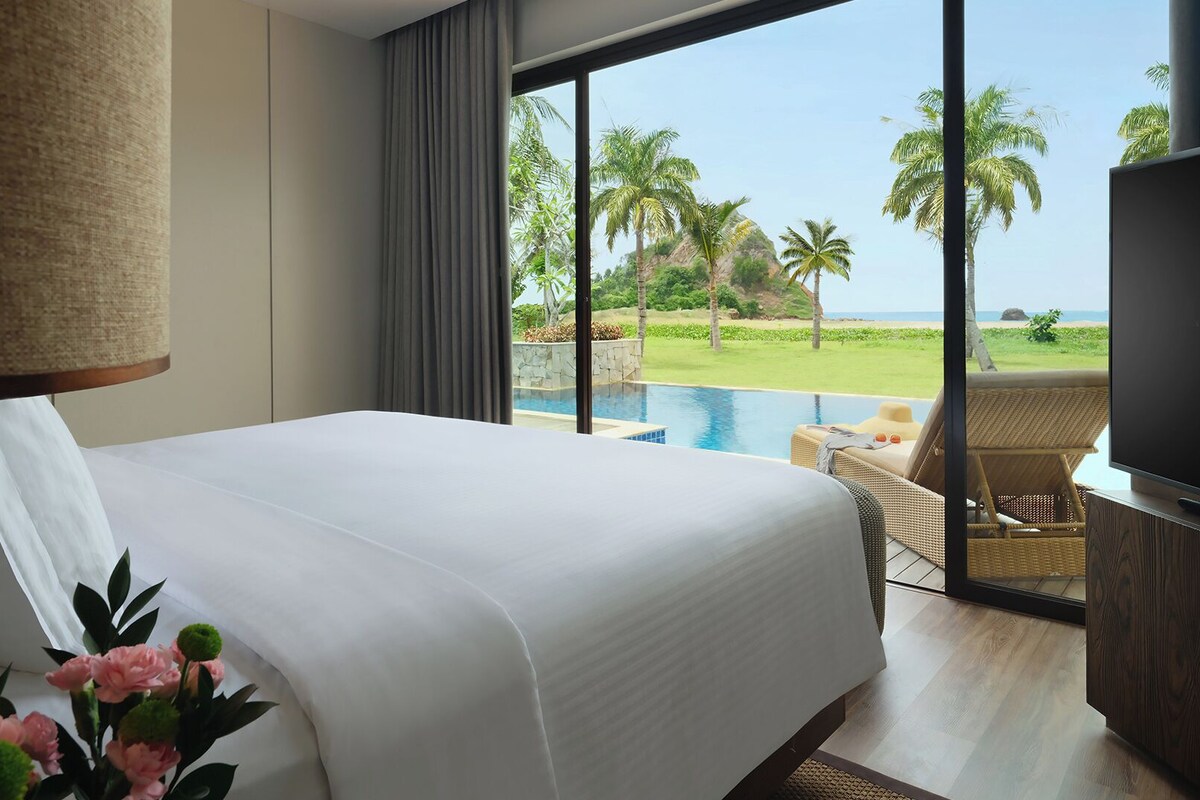 Een hotelkamer met een groot bed, een televisie, uitzicht op een zwembad en palmbomen.