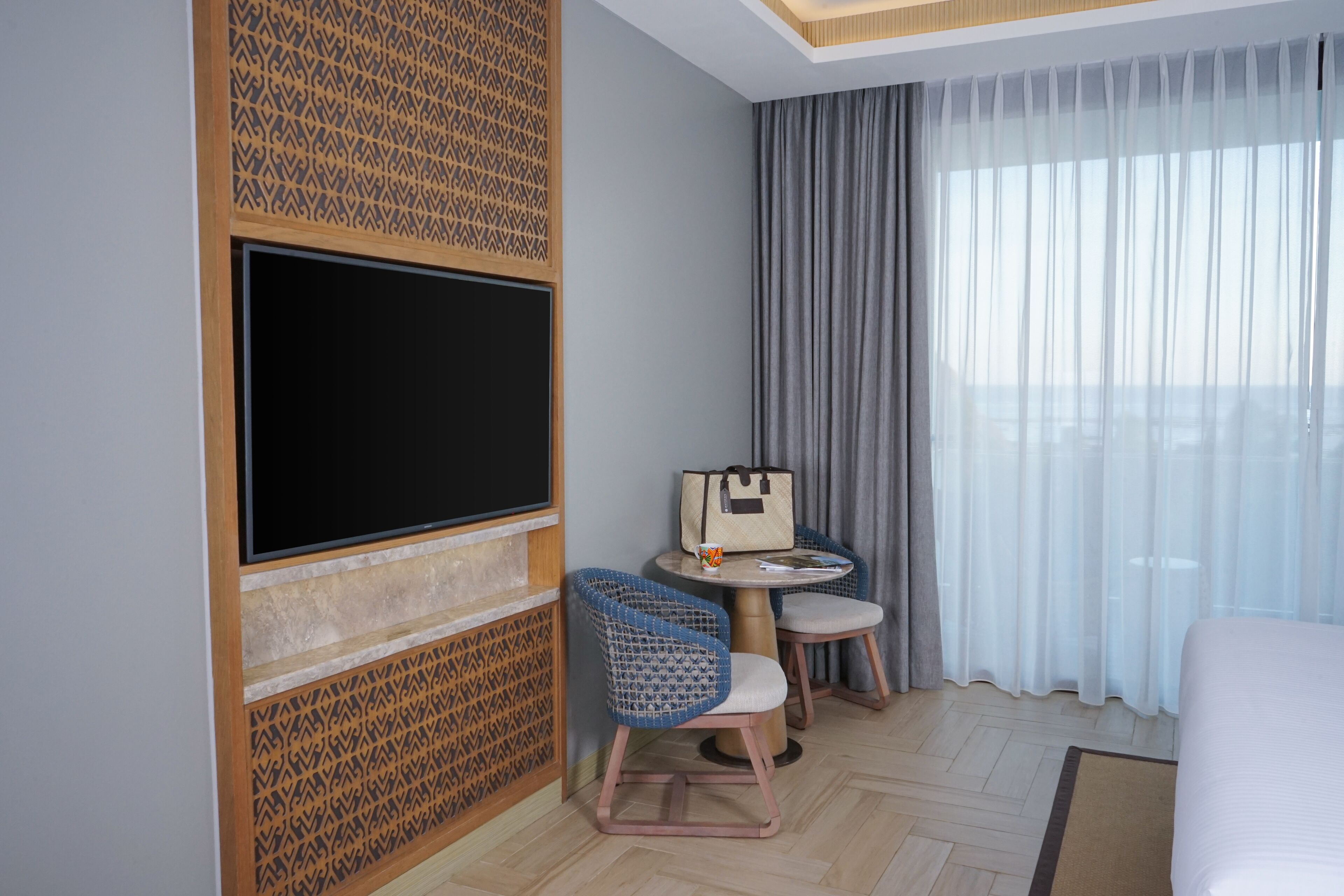 A hotel room with a large flat-screen TV, a small round table with a chair, a bed, and a window with sheer curtains.