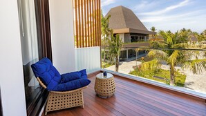 Un balcon avec un fauteuil en osier et une table ronde, donnant sur une plage bordée de palmiers et sur un bâtiment moderne surmonté d’un toit en forme de pyramide.