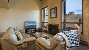 Ein Wohnzimmer mit Ledersofas, einem Flachbildfernseher, einem Kamin und großen Fenstern mit Blick auf die Berge.