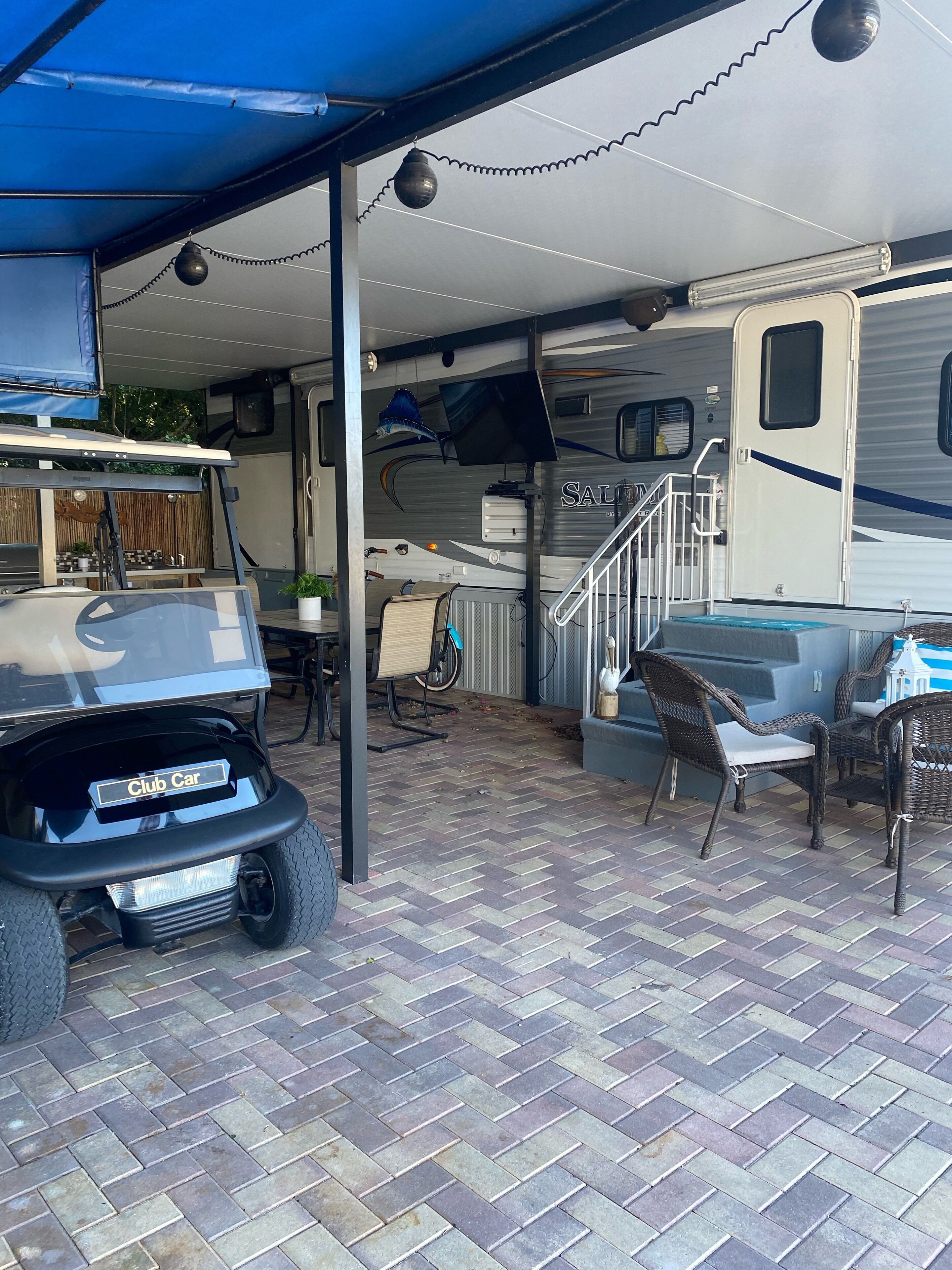 Un espacio al aire libre cubierto con un carro Club Car, una mesa de comedor con sillas y una autocaravana grande con una escalera que lleva a una terraza.