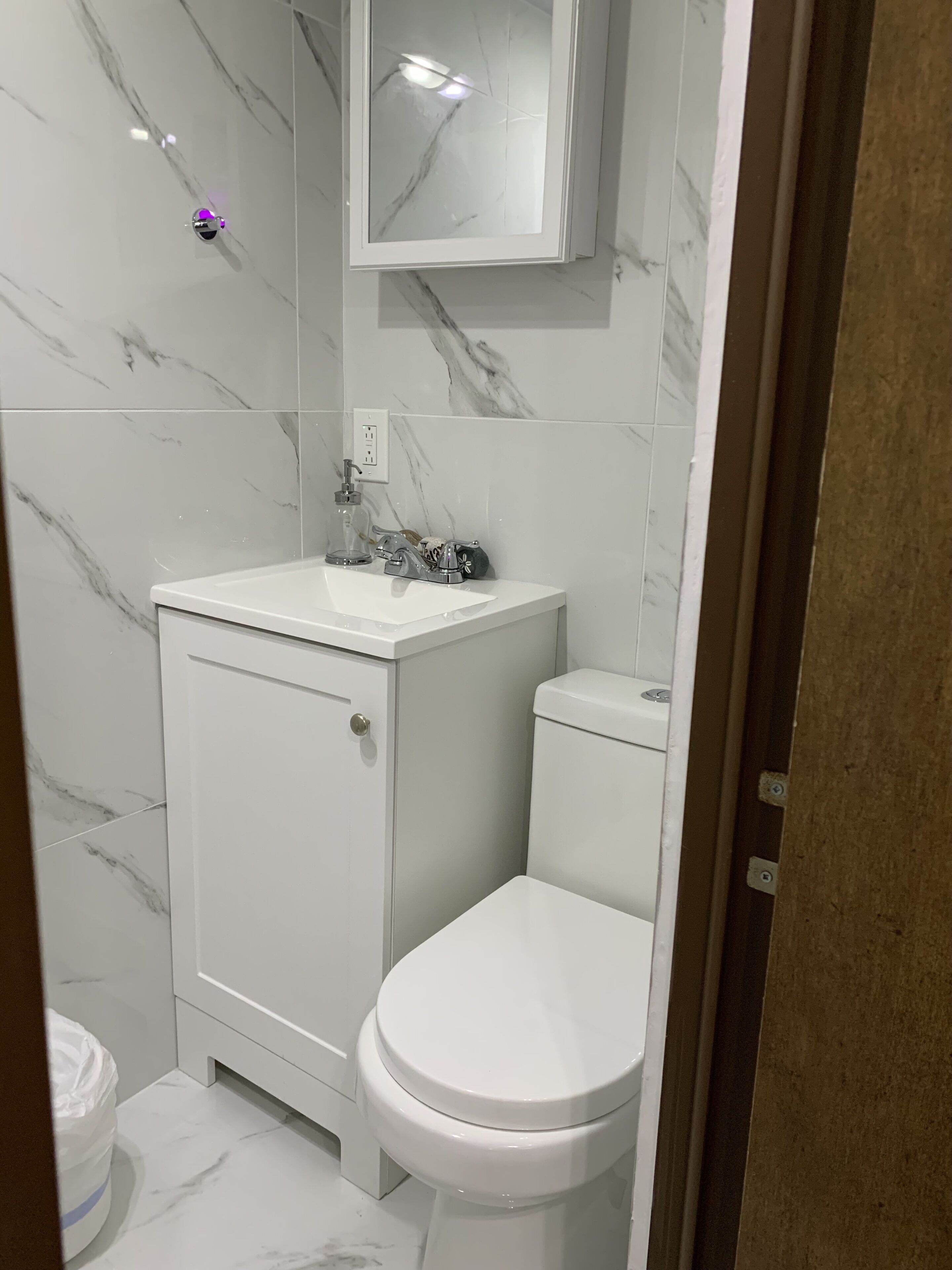 Ein modernes Badezimmer mit weißer Marmor-Spritzschutzwand, einem weißen Waschbecken mit Glas-Seifenspender, einer Toilette und einem Spiegel über dem Waschbecken.