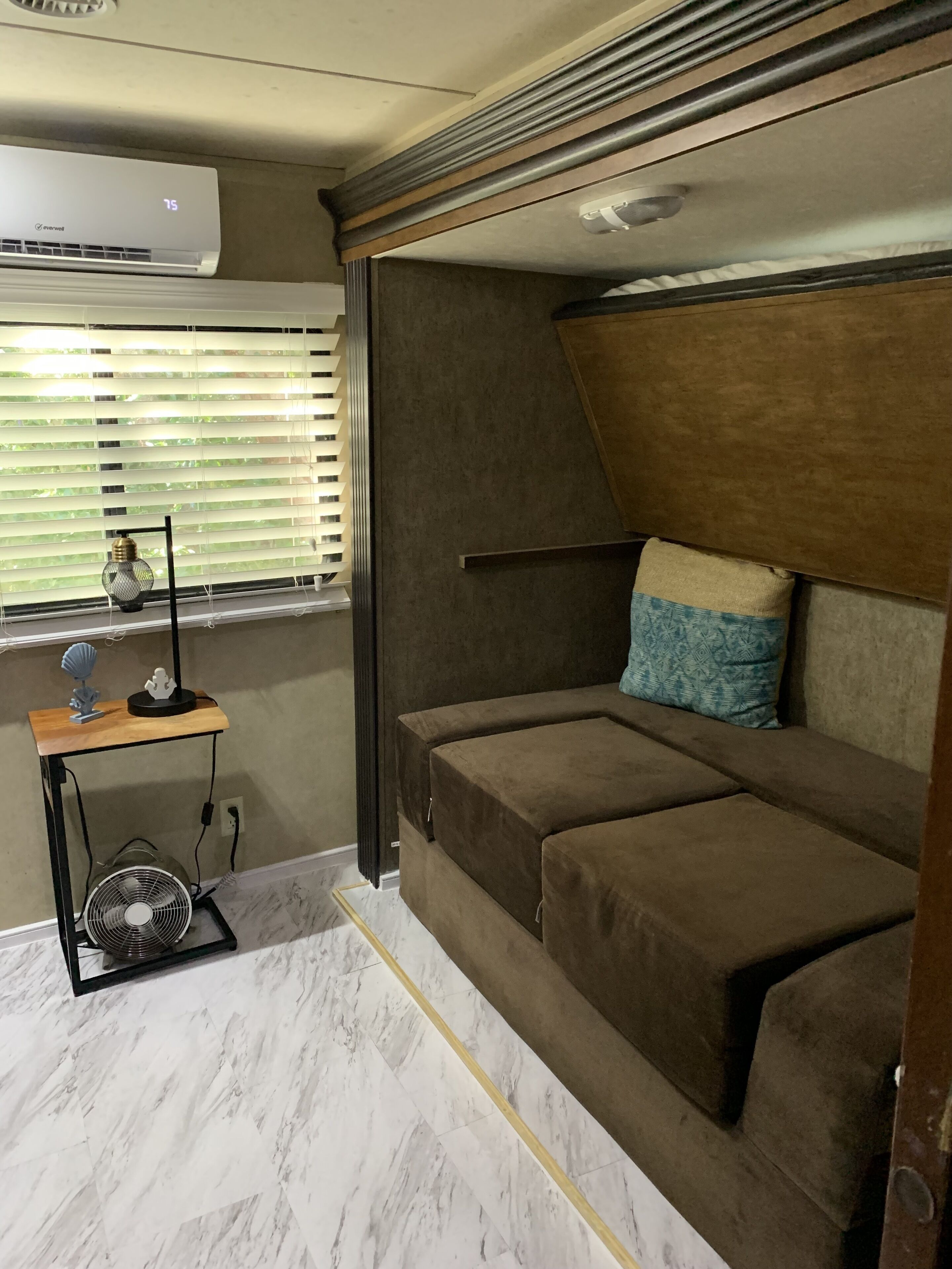 A compact bedroom with a sofa bed, a small table with a lamp, and a fan on the floor.