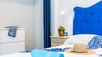 A neatly made bed with a blue headboard, a rolled blue towel, a beige hat, and a small potted plant on a bedside table.