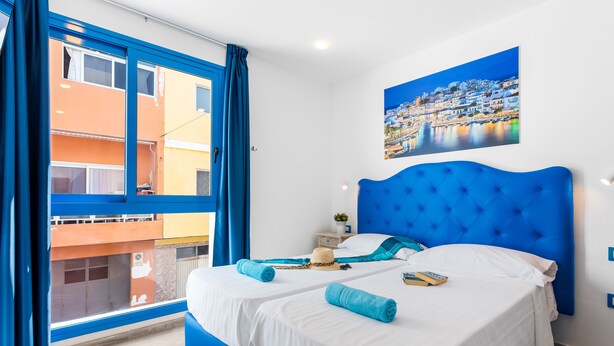 Un llit doble amb roba de llit blanca i blava, un panell frontal blau i vista d'un edifici a través d'una finestra amb cortines blaves.