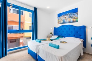 Een tweepersoonsbed met wit- en blauw beddengoed, een blauw hoofdeinde en uitzicht op een gebouw door een raam met blauwe gordijnen.