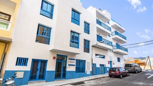 Un edifici de diversos pisos amb finestres i balcons amb marc blau, una porta amb el nom "OKEANOS" visible i cotxes aparcats al carrer.