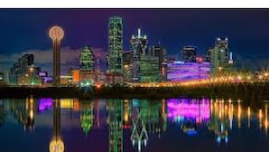 Une vue nocturne d’une ville, avec des bâtiments illuminés se reflétant dans une étendue d’eau.
