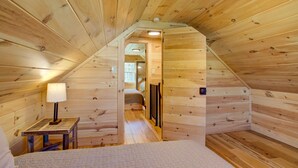 Un intérieur en bois avec un plafond en pente, un lit recouvert d’une courtepointe, une table de chevet avec une lampe et une porte donnant sur une autre pièce.