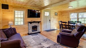 L’intérieur chaleureux d’un chalet avec une cheminée en pierre, des murs en bois, un téléviseur à écran plat, un coin repas avec des chaises et un canapé.