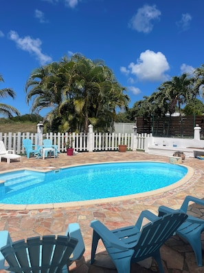 A swimming pool with a stone patio, surrounded by white picket fence, blue chairs, and palm trees.
