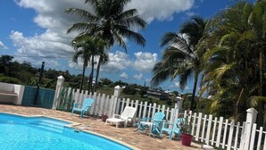 A swimming pool with a stone-tiled deck, surrounded by palm trees and a white picket fence.