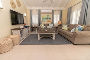A living room with a sofa, coffee table, and ottomans, a flat-screen TV, and a painting on the wall. - Paradise Villa|Gated|Pool|Richmond (Ocho Rios)