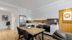 A modern kitchen with white cabinets, a stainless steel refrigerator, a built-in oven, and a dining area with a wooden table and chairs. - Mountain Views Penthouse - Apartment 22 (Queenstown)