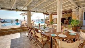 An outdoor dining area with a pool view, wooden furniture, and stone walls. - Villa Thalassa Thea (Corfu)