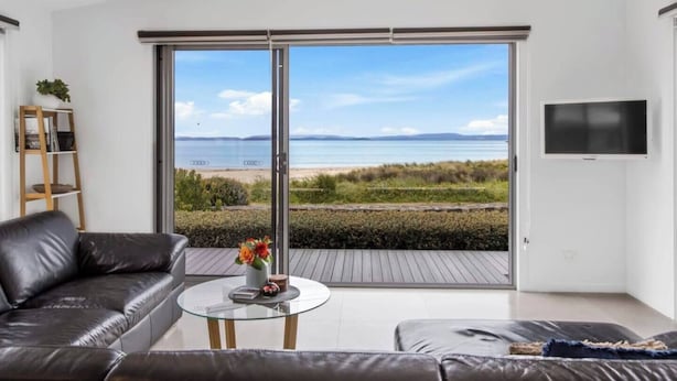 A modern living room with a large sliding glass door leading to a beach view, a leather sofa, a glass coffee table, and a TV mounted on the wall. - Seven Mile Beach House Surf Hike Golf (Seven Mile Beach)
