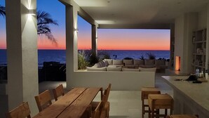 Ein moderner Wohnbereich mit Meerblick bei Sonnenuntergang, mit einem Holztisch, Sitzgelegenheiten und einem Bücherregal.