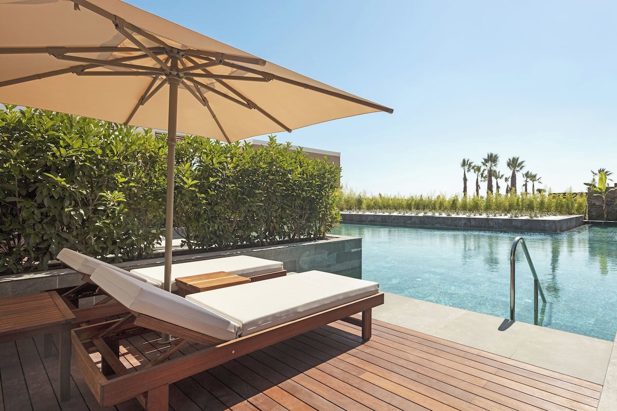 A poolside area with wooden lounge chairs and a large umbrella, surrounded by greenery and a clear pool.