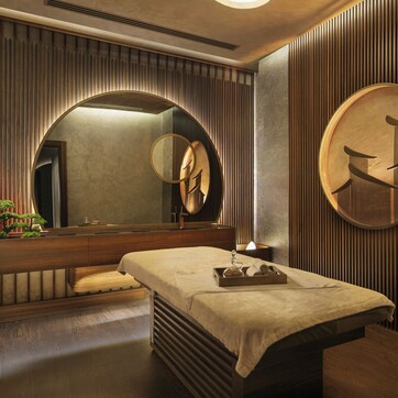 A modern spa room with a wooden bench, a massage table, a large mirror, and a wall-mounted towel rack.