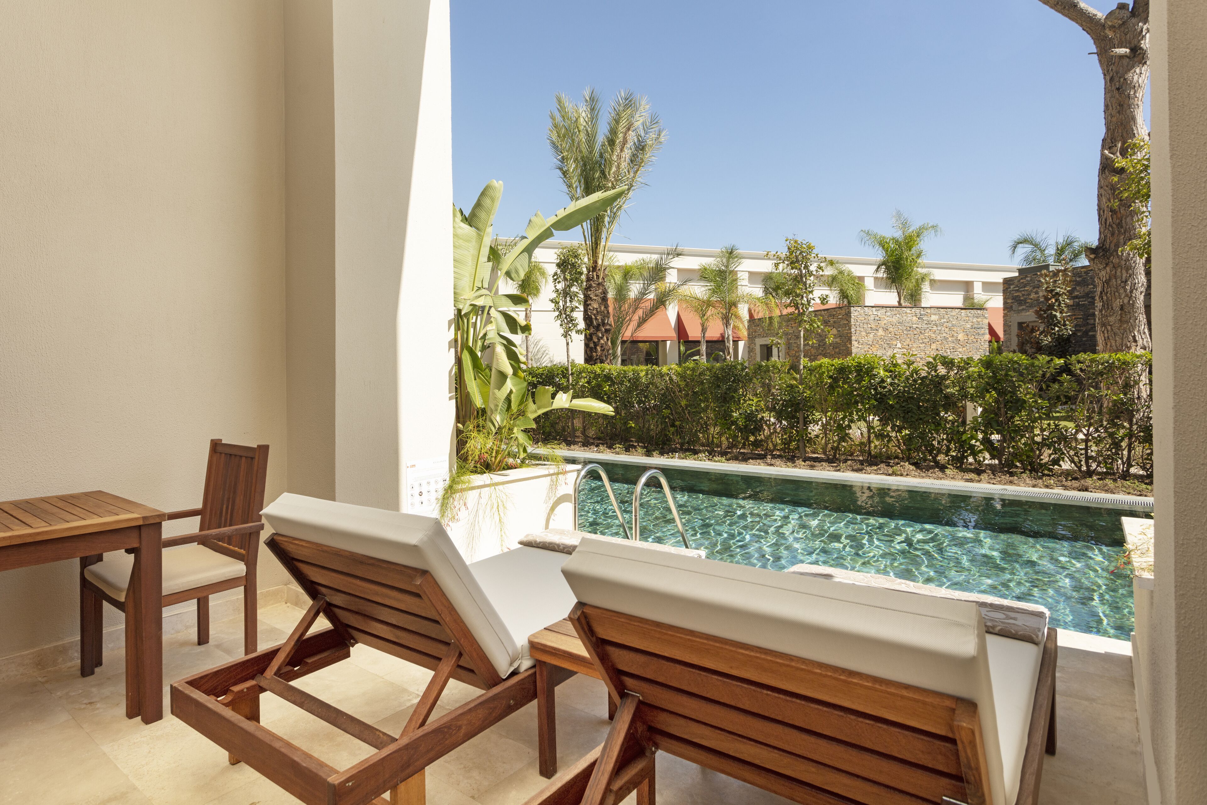 A poolside area with wooden lounge chairs and a table, a clear view of a swimming pool, and a building with a red roof in the background.