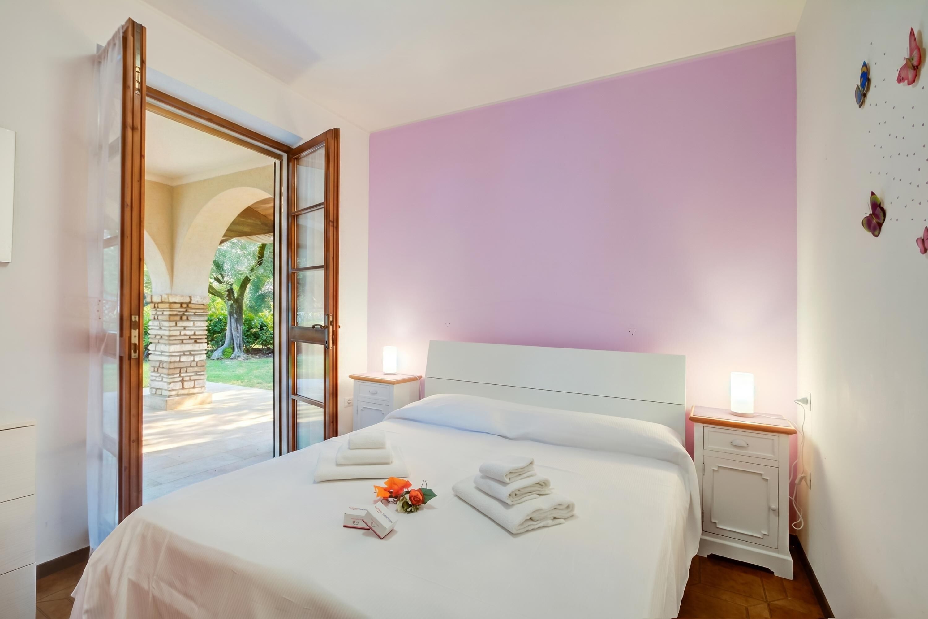 Ein Schlafzimmer mit einem Bett, zwei Nachttischen, einem Fenster und einer offenen Tür, die auf eine Terrasse führt.