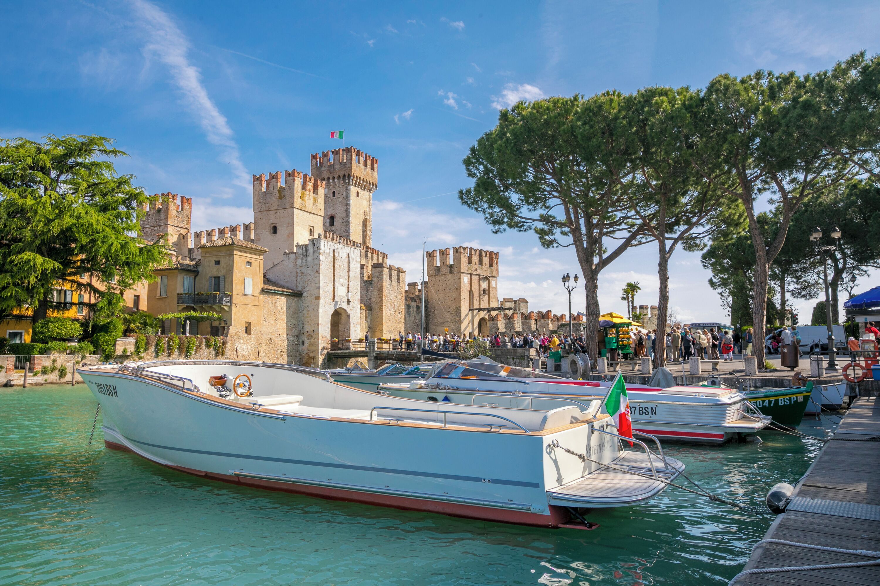 A historic castle with a moat, boats docked in the water, and a crowd of people in the background.