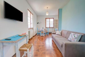 A living room with a sofa, a television mounted on the wall, a dining table with chairs, and a kitchen area with a window.