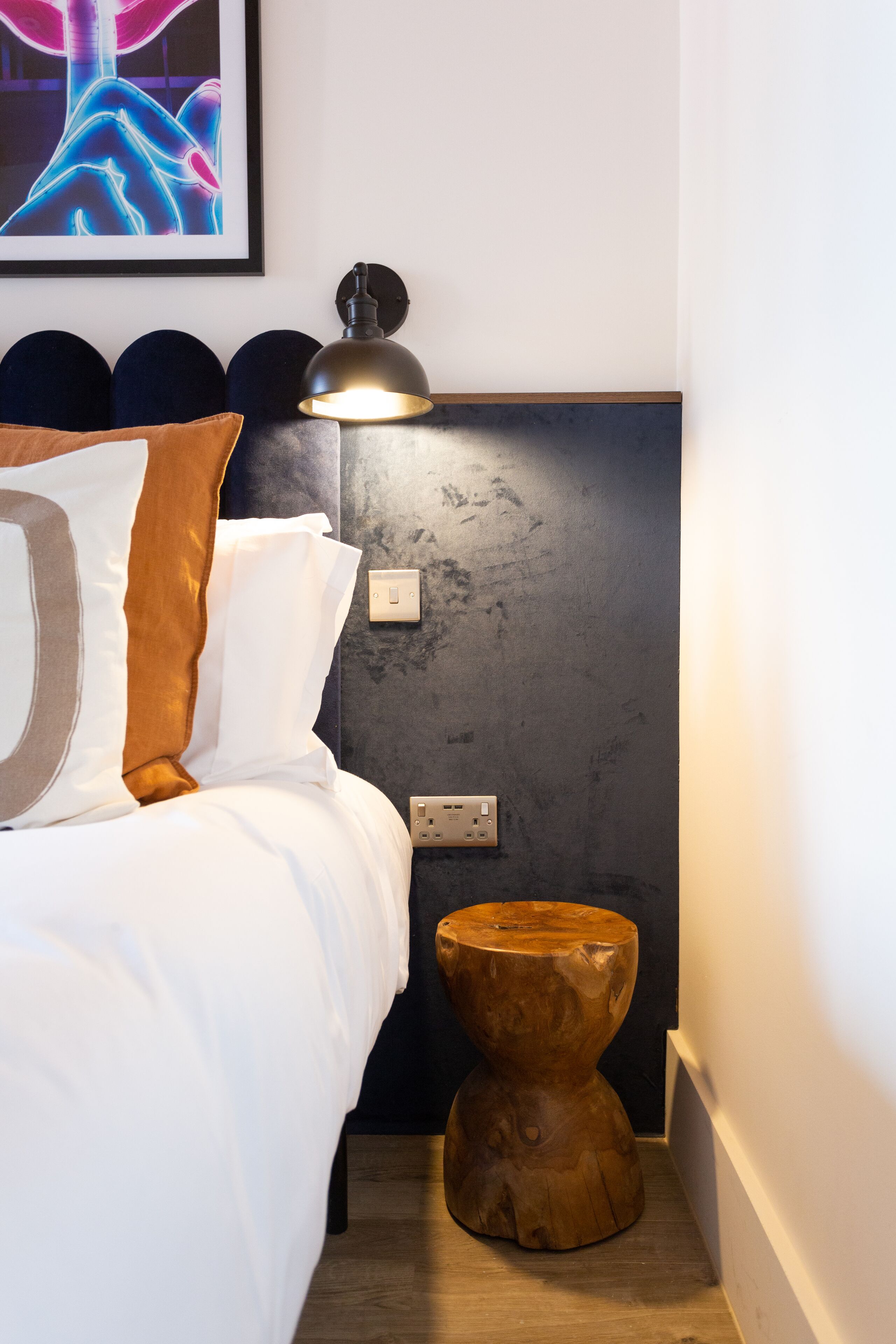 A neatly made bed with white and brown bedding, a wooden nightstand with a lamp, and a framed abstract artwork above the bed.
