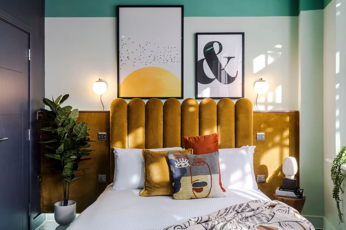 A neatly arranged bedroom with a wooden headboard, a large bed with patterned bedding, two wall-mounted reading lamps, and a framed artwork above the bed.