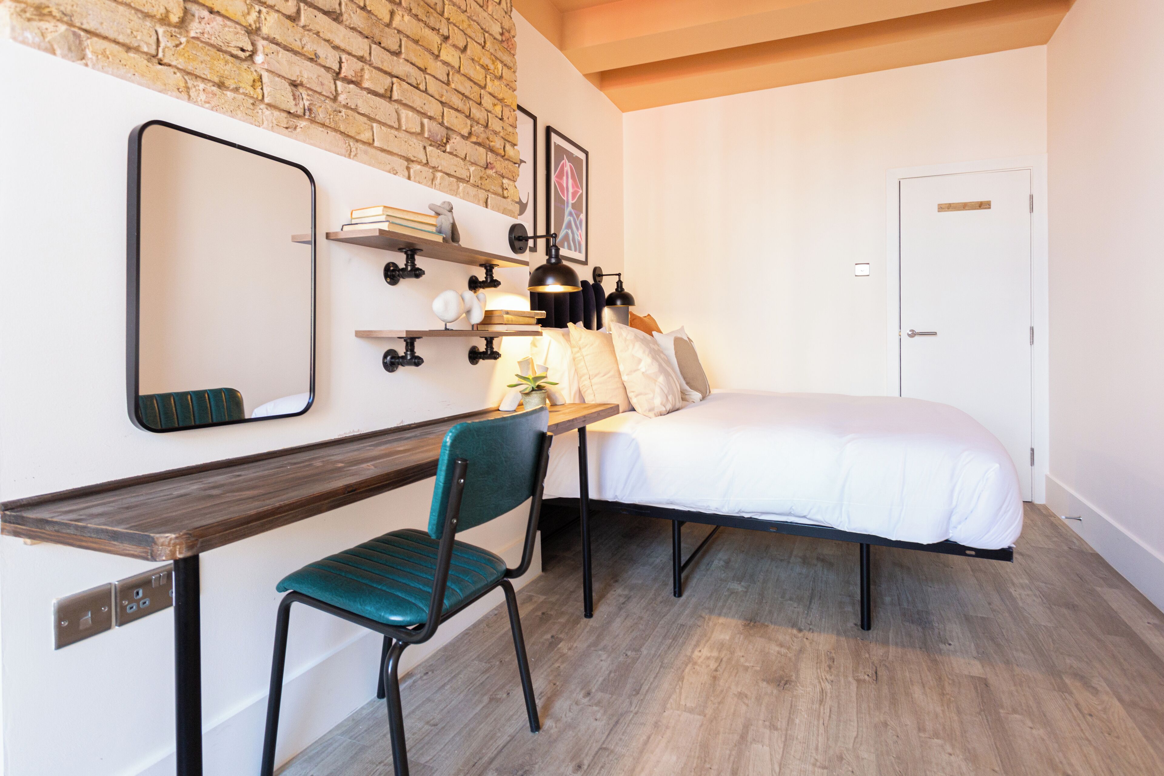 A bedroom with a bed, a desk with a chair, a mirror, shelves with books and decorative items, and a door.