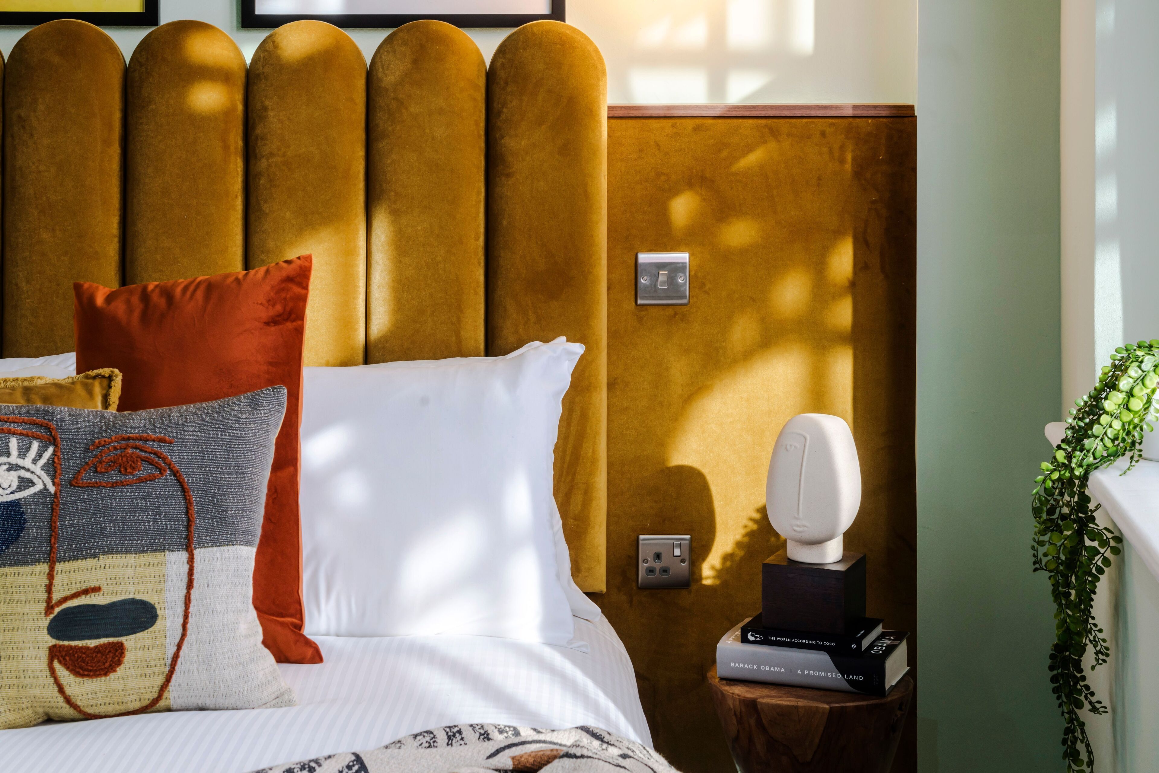 Un lit bien fait, agrémenté de plusieurs coussins, une table de chevet avec une lampe et des livres, et un interrupteur fixé au mur.
