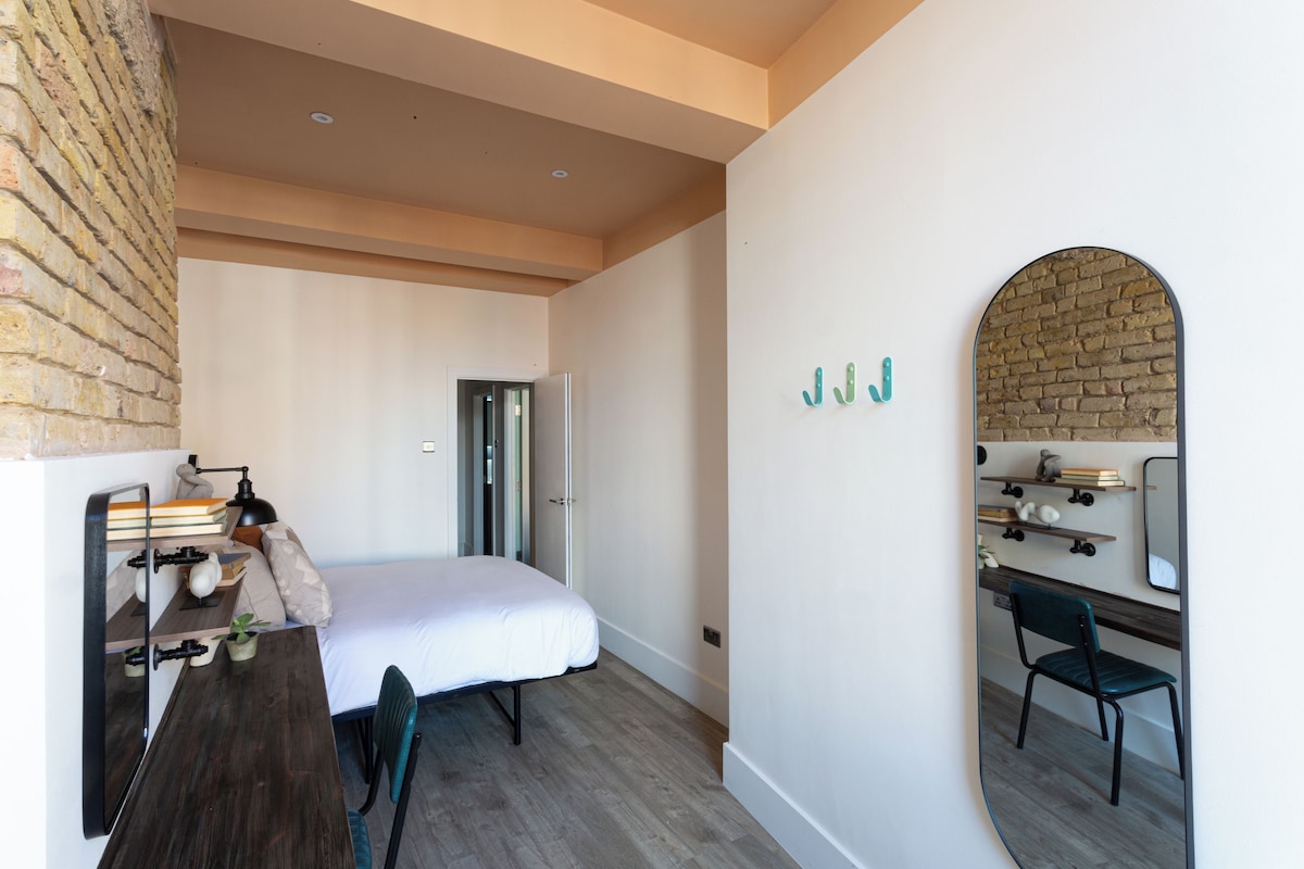 A bedroom with a bed, a desk, a chair, a mirror, and a brick wall.