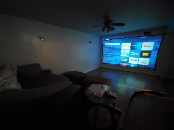 A dimly lit room with a large screen displaying a TV menu. There is a ceiling fan, a couch, and a small table with a projector.