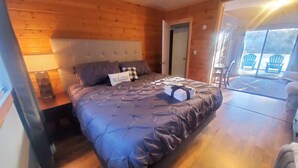 Une chambre à coucher aux murs lambrés, avec un grand lit, une table de chevet, une commode et une vue sur l’extérieur grâce à une porte vitrée.
