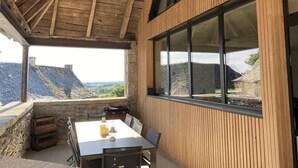 Ein überdachter Essbereich im Freien mit Tisch, Stühlen, Grill und Blick auf die Landschaft.