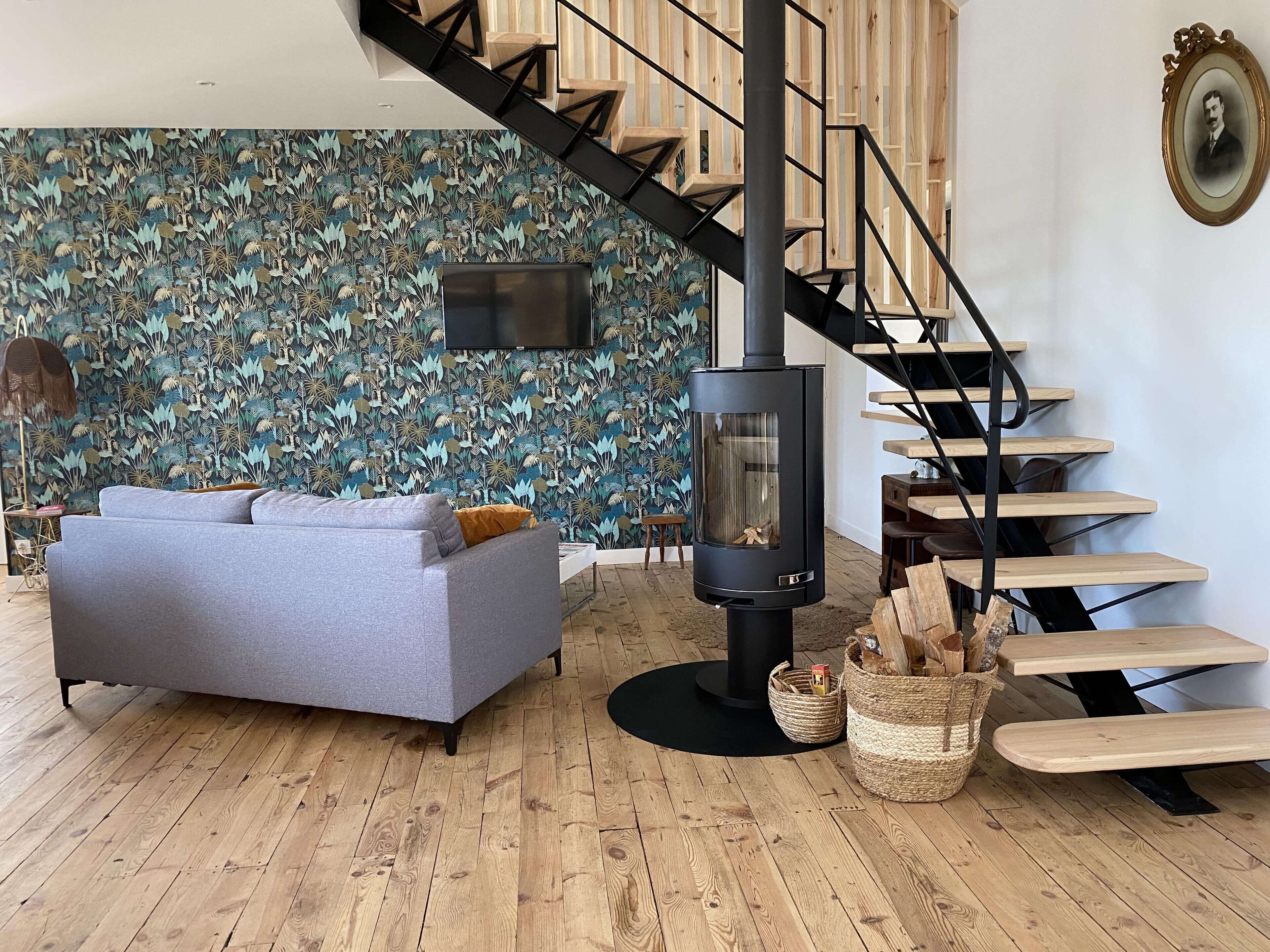 A modern living room with a grey sofa, a black wood-burning stove, a wooden staircase, and a patterned wall.