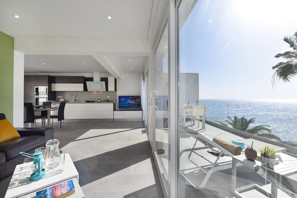 A modern living space with a balcony overlooking the sea, a kitchen area, and a dining setup.