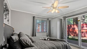 Un dormitorio con cama, ventilador de techo, ventana con persianas y puerta corredera de vidrio que da a un espacio exterior.