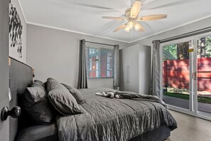 Una camera da letto con un letto, una ventola a soffitto, una finestra con persiane e una porta scorrevole in vetro che dà su uno spazio esterno.