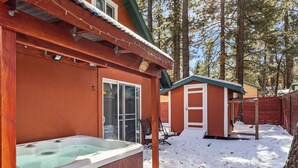 Una cabaña de madera con jacuzzi, un porche cubierto y un cobertizo pequeño en un bosque nevado.