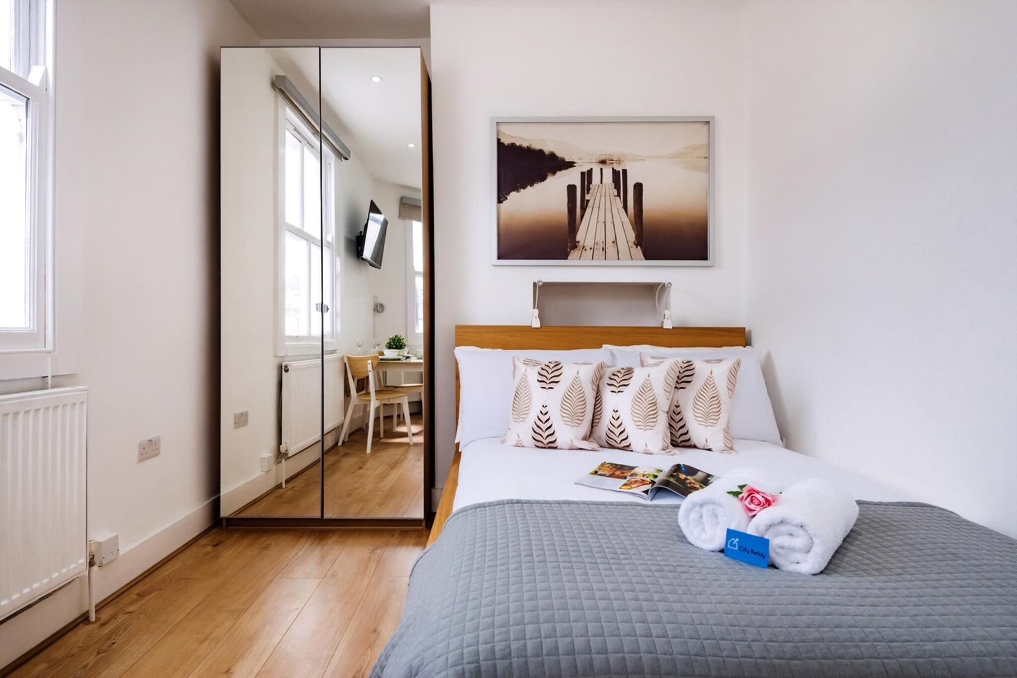 A hotel room with a bed, a painting above the bed, a desk, and a chair.