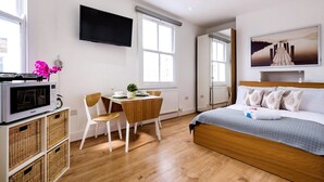 A modern hotel room with a wooden floor, a bed with a grey quilt, a flat-screen TV mounted on the wall, a dining table with chairs, and a small potted plant.