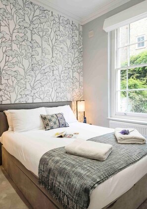 A bedroom with a bed, a nightstand, a lamp, a window with a view of greenery, and a patterned wallpaper.