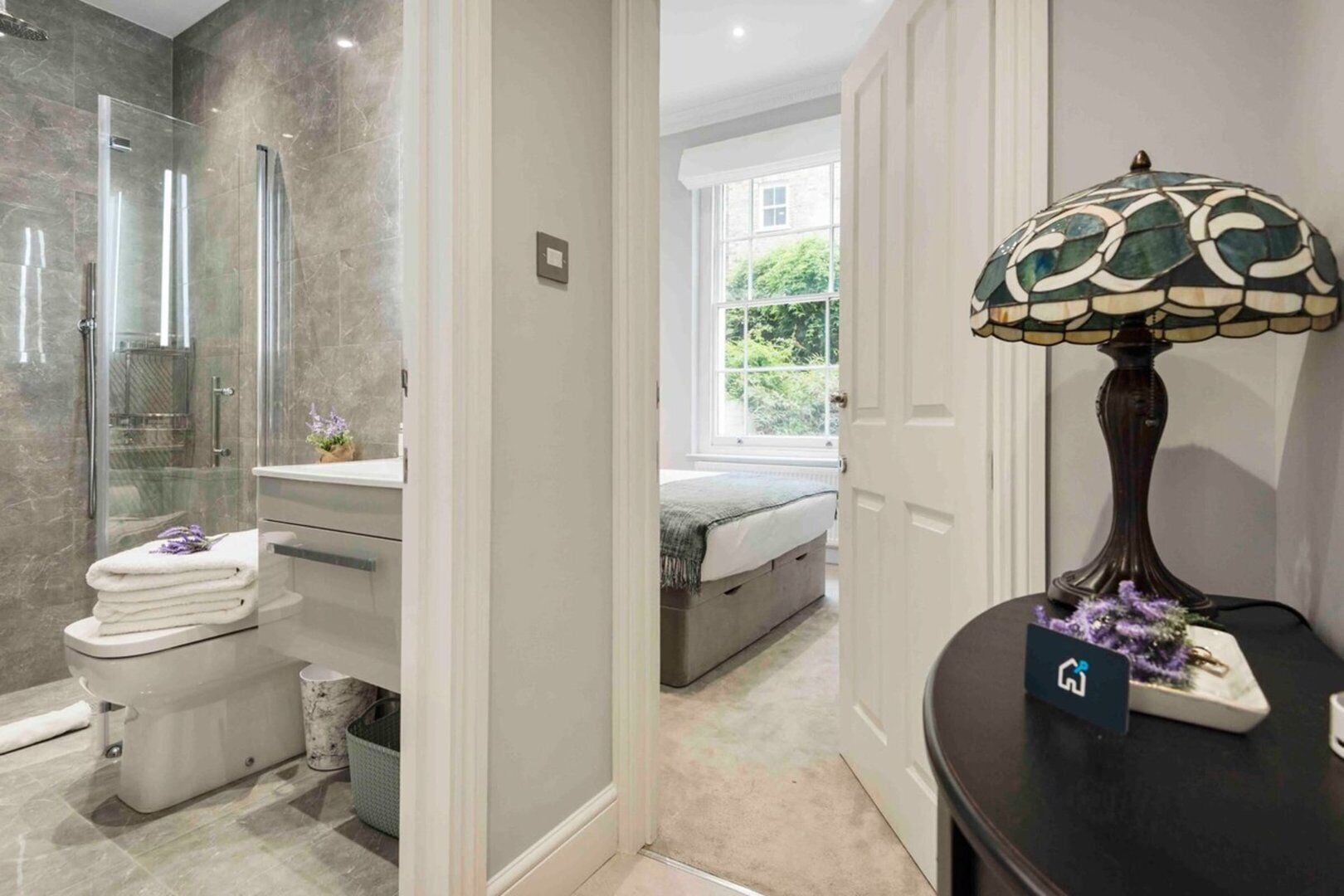 A modern bathroom with a glass shower, a toilet with stacked towels, and a bedside table with a lamp.