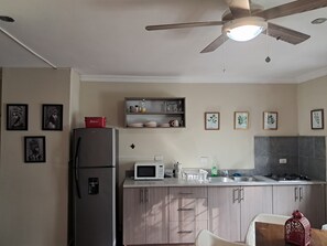 A kitchen with a refrigerator, microwave, sink, and cabinets. There are framed pictures on the wall and a ceiling fan.