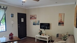 Un salon équipé d’un ventilateur de plafond, d’un téléviseur à écran plat, d’un canapé, d’une petite table avec une plante et d’une porte sur laquelle est apposée une pancarte.