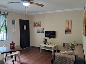Una sala de estar con ventilador de techo, televisor de pantalla plana, sofá, una mesita con una planta y una puerta con un letrero.