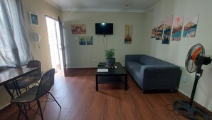 Una sala de estar con un sofá, una mesa de comedor con sillas, un televisor montado en la pared y una planta en maceta sobre una mesita auxiliar.