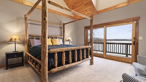 Una cama con dosel de madera, suelo alfombrado, una mesita de noche con lámpara y vistas a un balcón con barandilla.