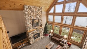 Un salón acogedor con chimenea de piedra, un televisor grande y vistas al exterior a través de amplios ventanales.